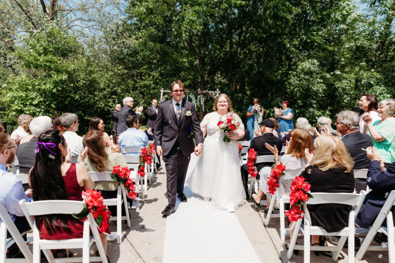 Bride and groom walk down an outdoor wedding aisle lined with white chairs decorated with red flowers, as guests look on.