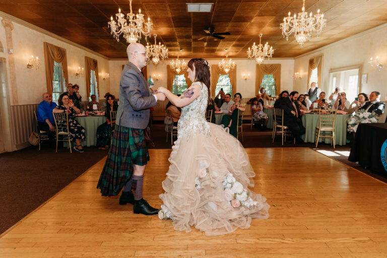Bride in a voluminous white wedding gown dances with a groom wearing a kilt on a wooden dance floor, while guests sit at tables in a chandelier-lit ballroom.