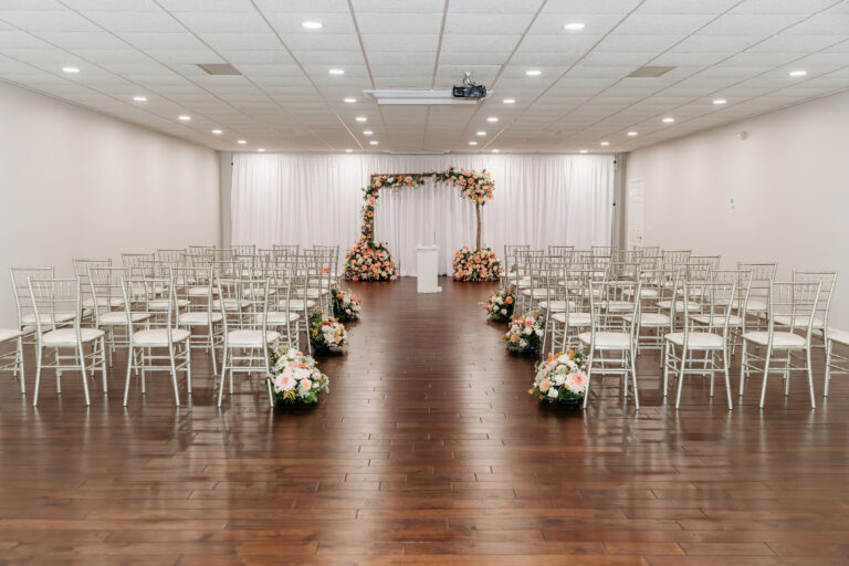 Rows of white chairs line a wooden aisle toward a floral arch at the front of a bright ceremony space.