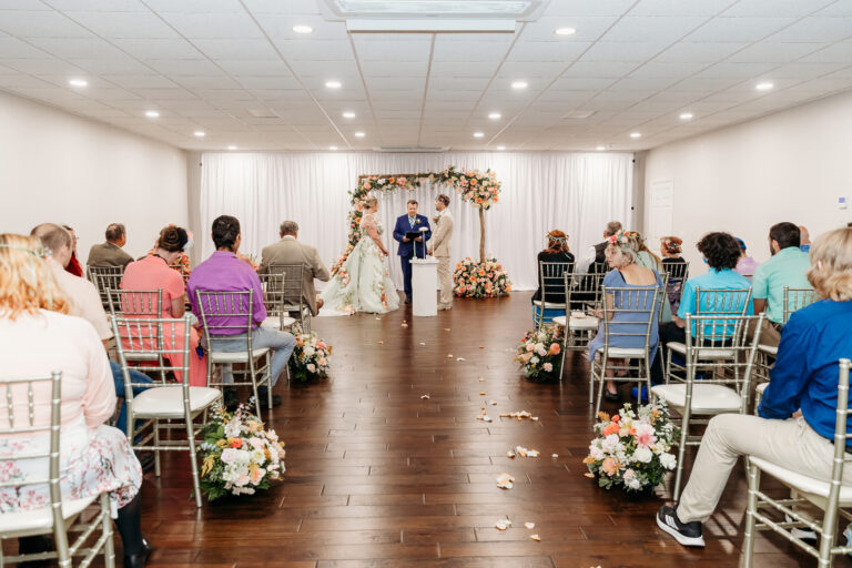Indoor wedding ceremony with a couple standing under a floral arch while guests sit in rows of chairs.