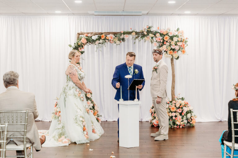 Bride in a floral gown and groom in a light suit stand with a blue-suited officiant at a white podium under a flower-filled arch as guests watch the ceremony.