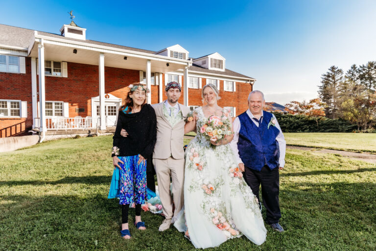 Bride in a floral-embroidered white wedding dress poses with guests on a sunny lawn in front of a red brick building.