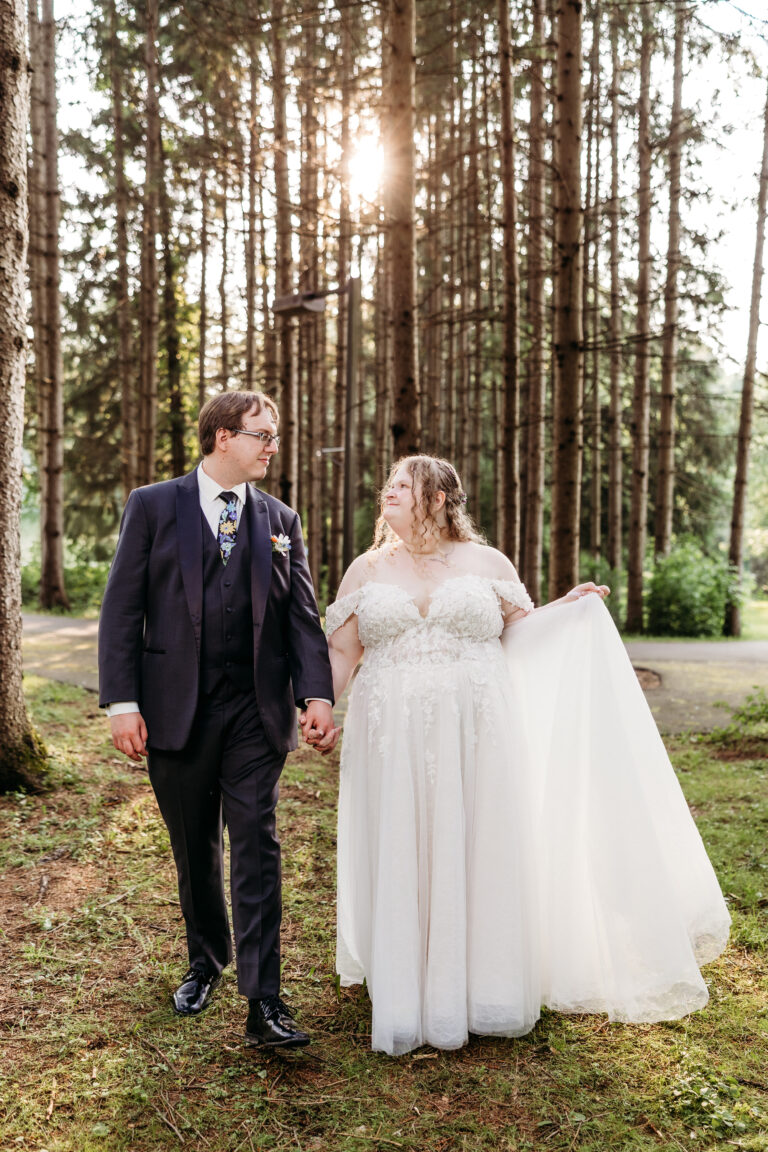 Bride in a white wedding gown and veil and a groom in a dark suit walk hand in hand through a sunlit forest.