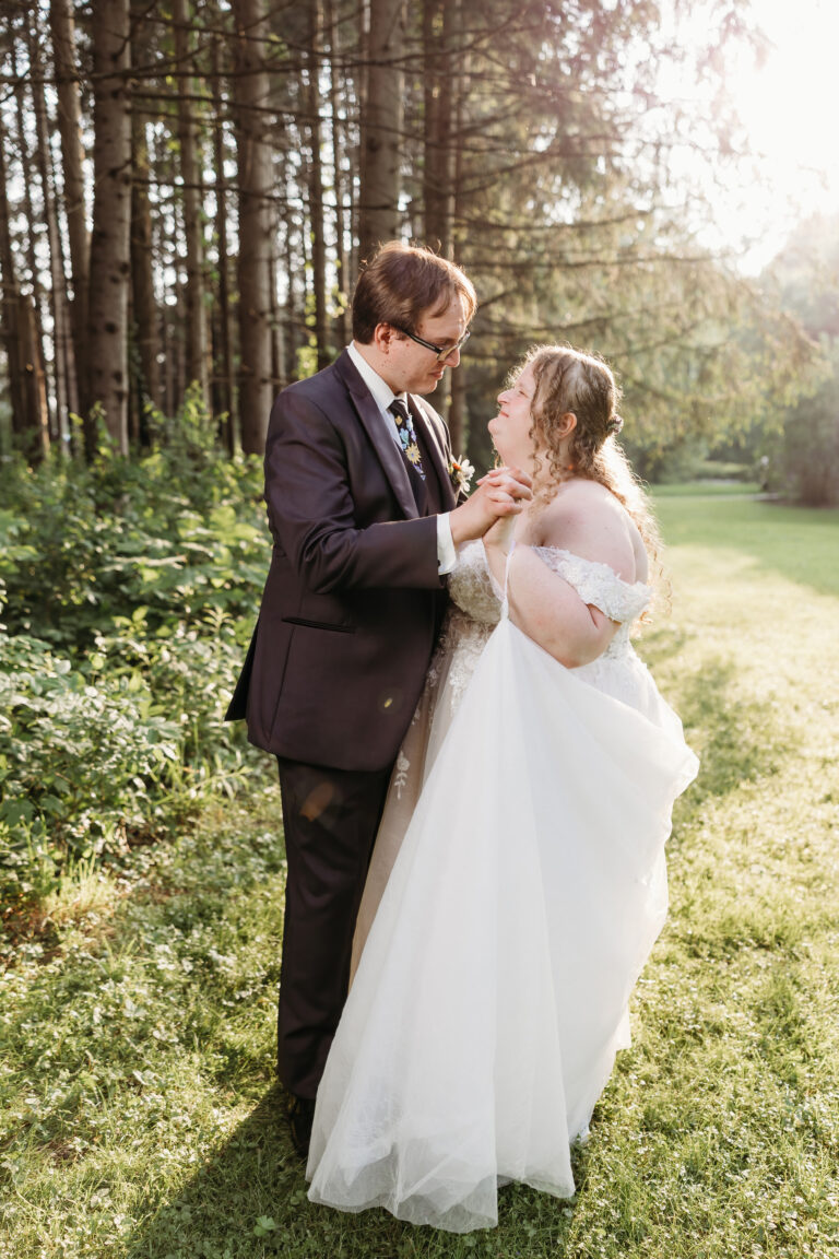 A bride in a white wedding gown and a groom in a dark suit stand in a sunlit meadow, gazing at each other.