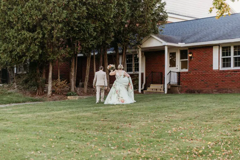 A man in a tan suit and a woman in a floral wedding dress walk across a grassy lawn toward a brick house.