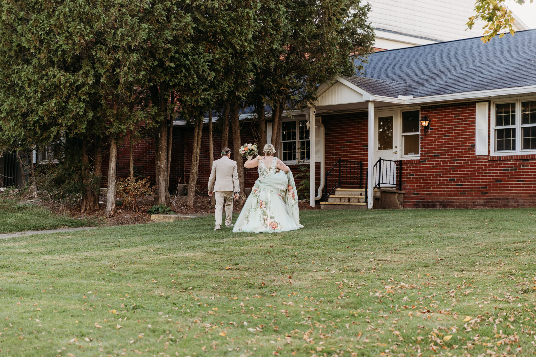 A man in a tan suit and a woman in a floral wedding dress walk across a grassy lawn toward a brick house.