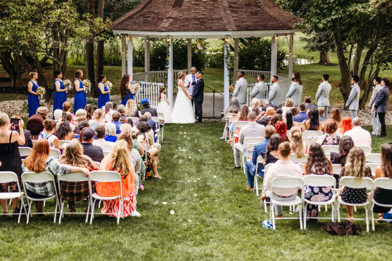 Outdoor wedding ceremony under a gazebo with the bride and groom kissing, flanked by bridesmaids in blue dresses and groomsmen in light suits, while guests sit on white chairs on the grassy lawn.