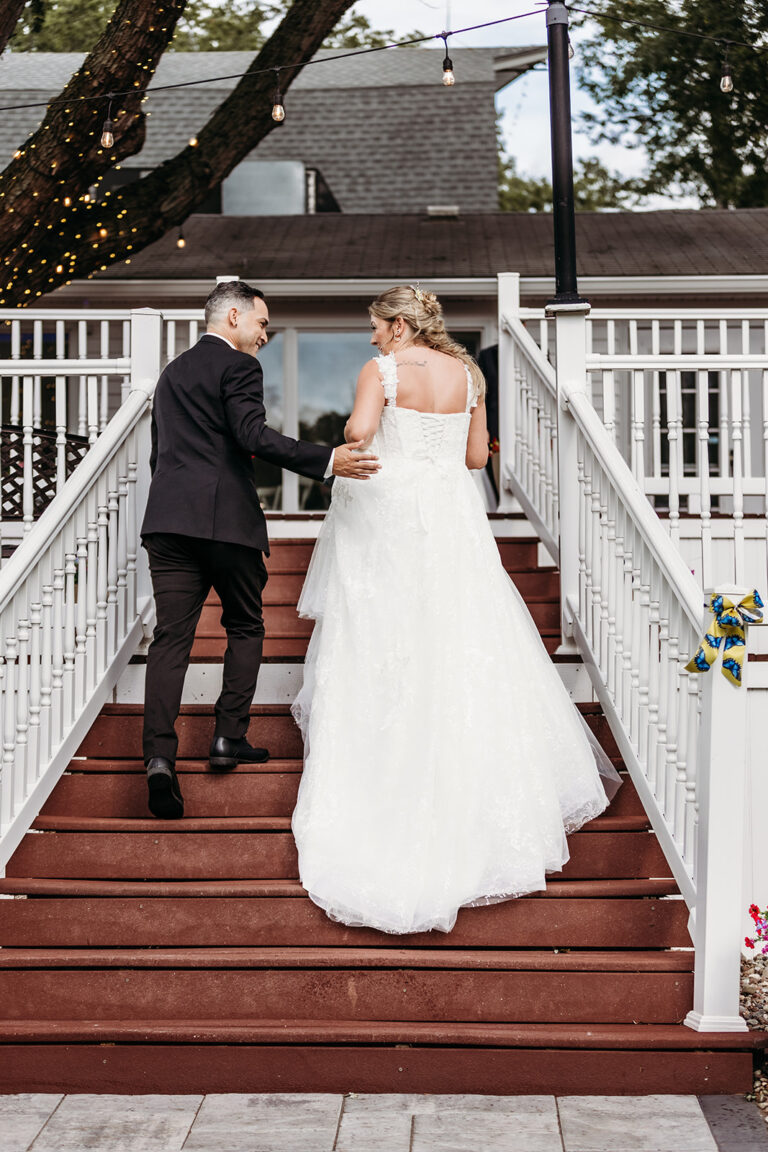 A bride in a white wedding dress and a groom in a dark suit walk up a wooden staircase at an outdoor wedding.