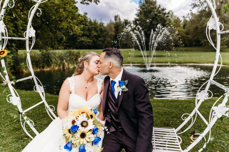 A bride and groom kiss on a decorative white garden swing beside a pond with a fountain.