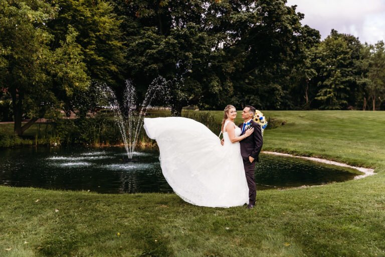 A bride in a white wedding gown and a groom in a dark suit embrace beside a pond with a fountain on a grassy lawn.
