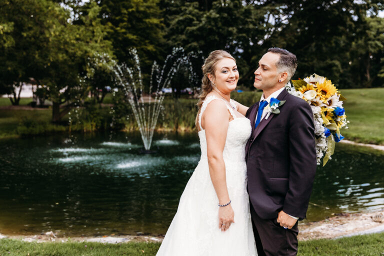 Bride in a white wedding dress and groom in a dark suit pose beside a pond with a fountain in a park.