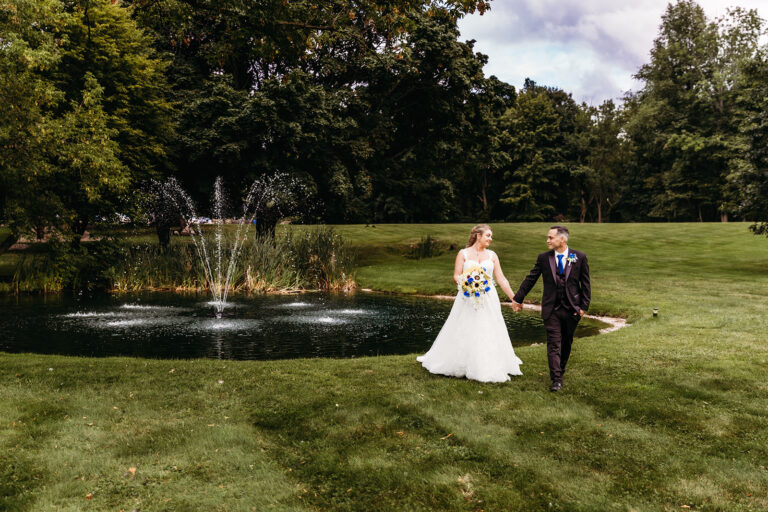 Bride in a white wedding gown and groom in a dark suit walk hand in hand across a grassy park beside a pond with a fountain.