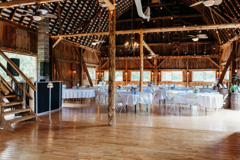 Rustic barn interior with wooden beams, string lights, and round tables set for a reception.