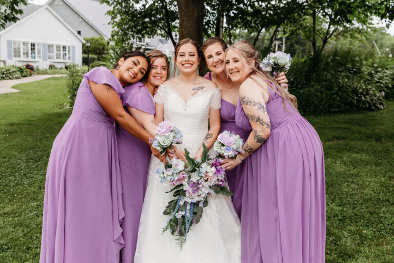 Bride in a white wedding gown stands outdoors with four bridesmaids in lavender dresses, all smiling and posing with the bride’s bouquet.
