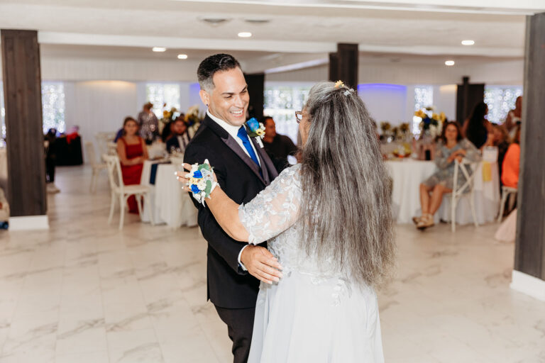 A bride and groom dance together at a bright wedding reception with guests seated at tables in the background.