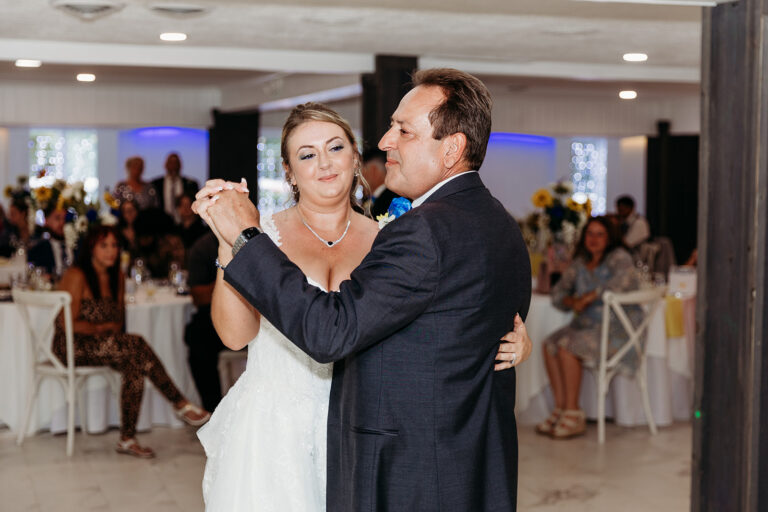 A couple dances closely at a wedding reception with guests seated at tables in the background.