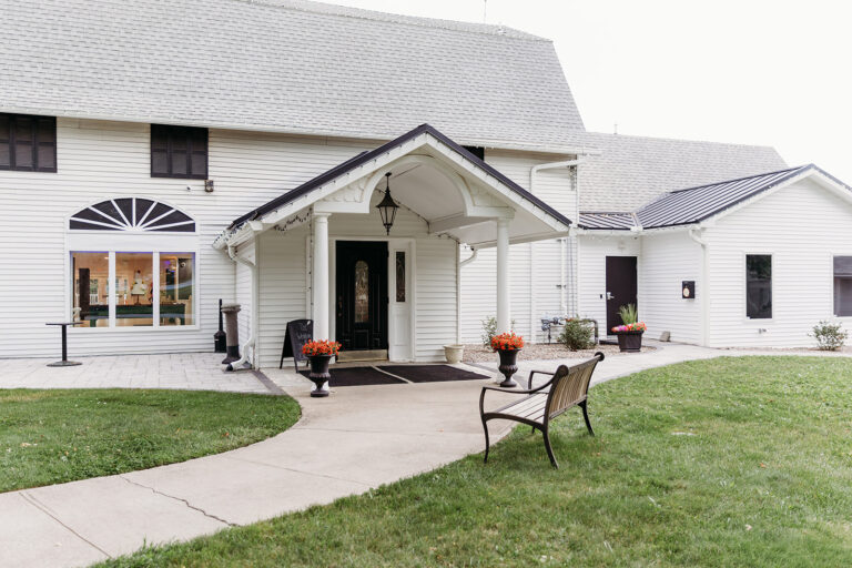 White building with a covered front porch, curved walkway, and a black bench beside red potted plants at the entrance.