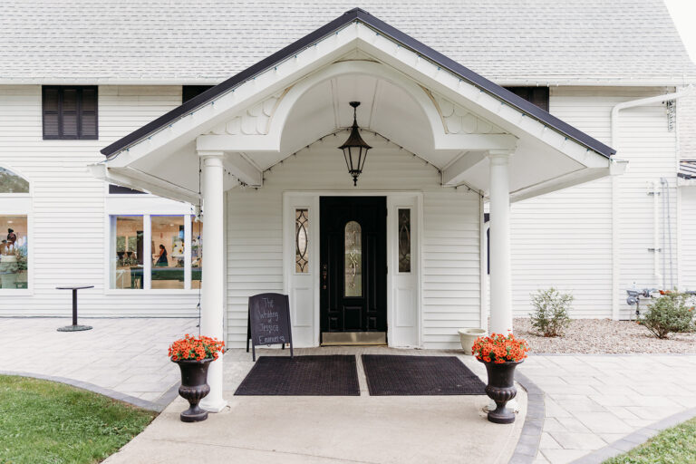 A white building with a gabled front porch and a black door flanked by two orange flower pots.