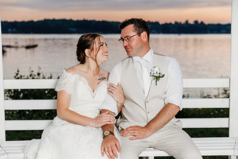 Bride in a white wedding dress and groom in a light suit sit close on a white bench by a sunset lake, smiling at each other.