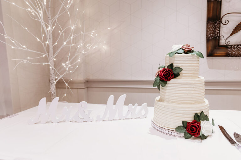 Three-tier white wedding cake adorned with red roses and green leaves, set on a white-draped table beside a lit decorative white tree.