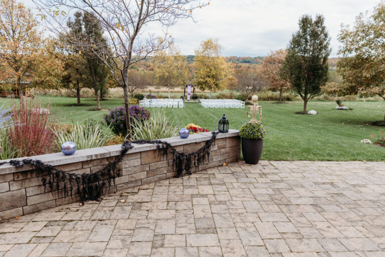 Stone-paved patio with a low brick wall lined with potted plants, overlooking a well-kept lawn with autumn-colored trees.
