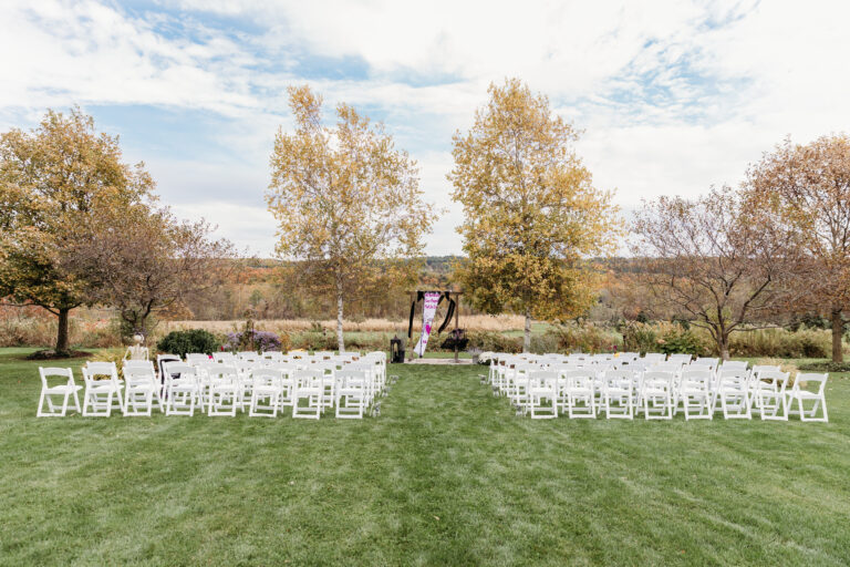 Outdoor wedding ceremony setup on a grassy lawn with white folding chairs in rows and a flower-draped arch at the far end.
