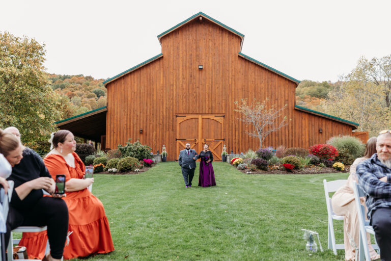 Outdoor wedding ceremony in front of a rustic wooden barn, with guests seated along a grassy aisle.
