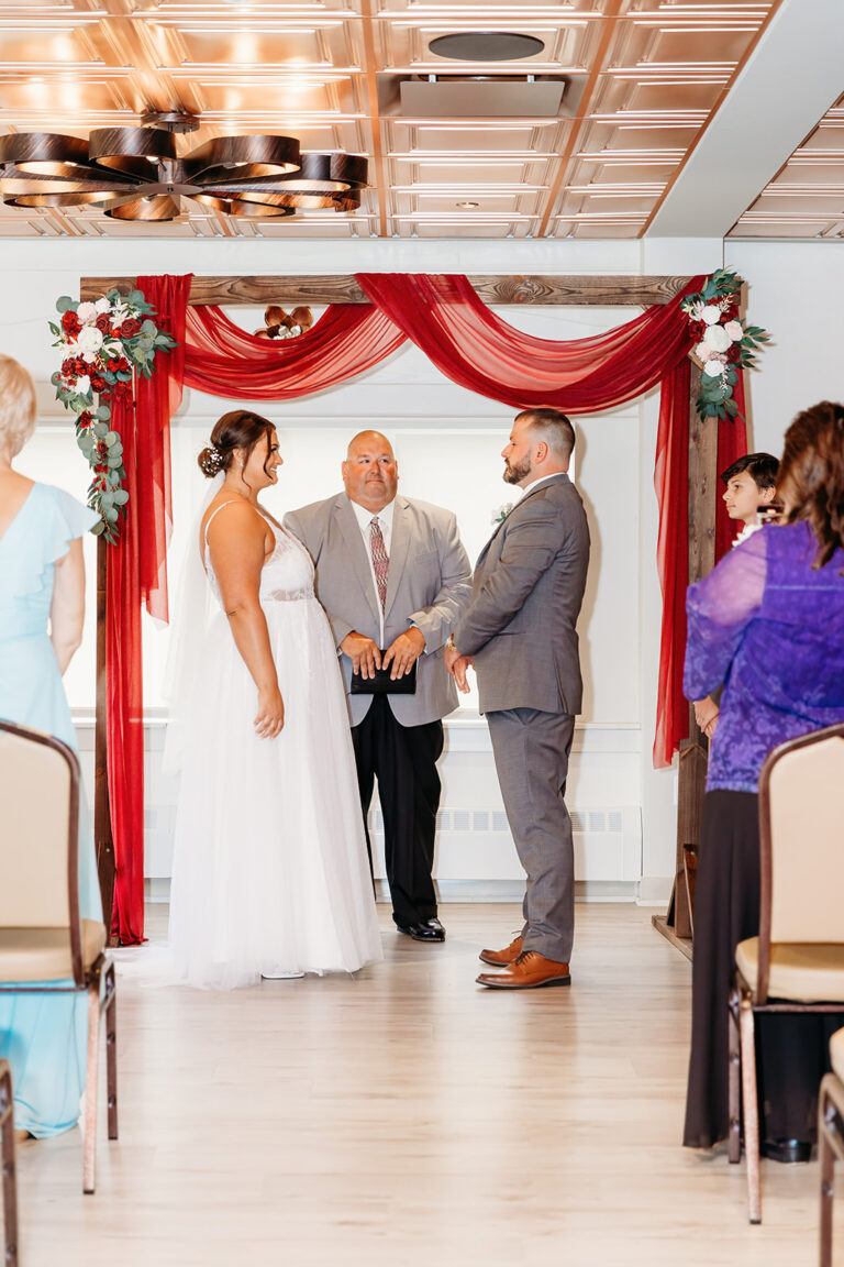 Bride and groom exchange vows with an officiant under a red floral arch as guests look on.