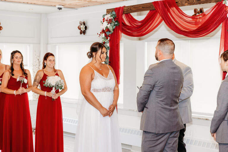Bride in a white wedding gown faces the groom in a gray suit during a ceremony, with bridesmaids in red dresses holding bouquets nearby.