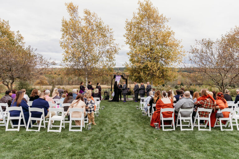 An outdoor wedding ceremony on a grassy lawn with rows of white folding chairs filled with guests, facing the officiant at the front under autumn trees.