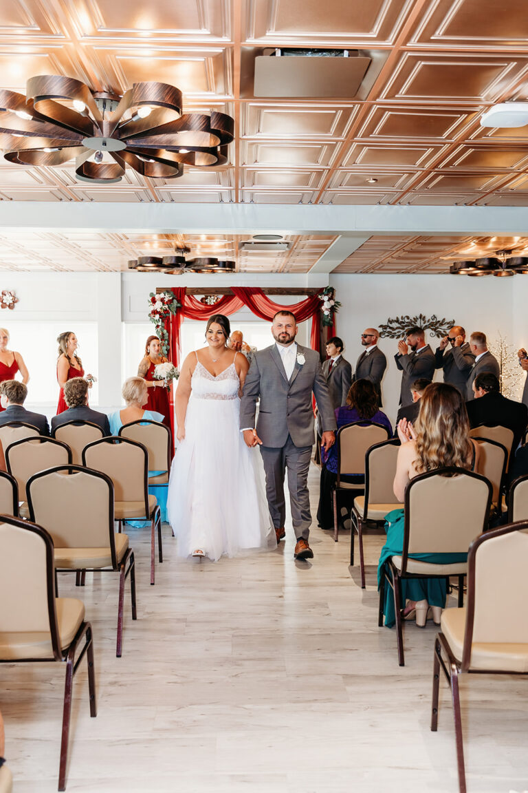 Bride in a white wedding dress and groom in a gray suit walk down the aisle at an indoor wedding ceremony with guests seated on both sides.