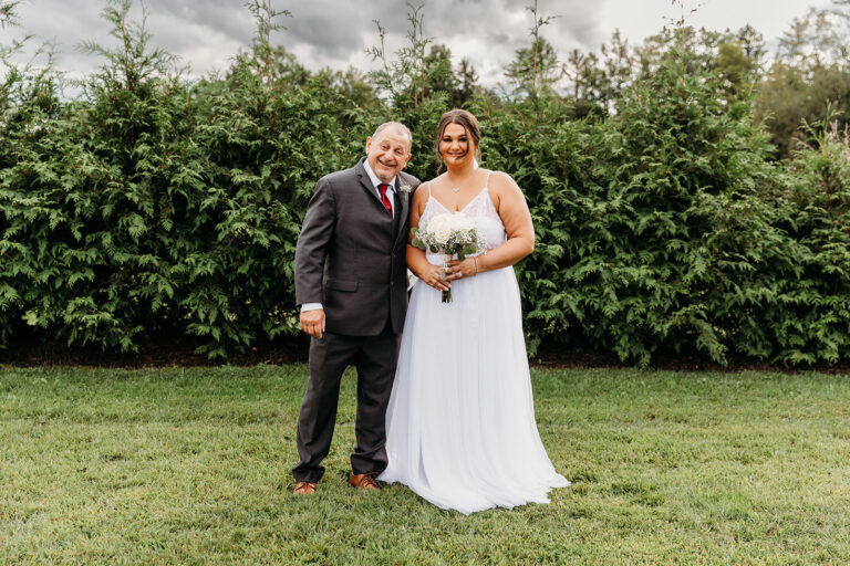 Bride in a white wedding gown holding a bouquet stands beside a man in a dark suit on a grassy lawn with trees in the background.