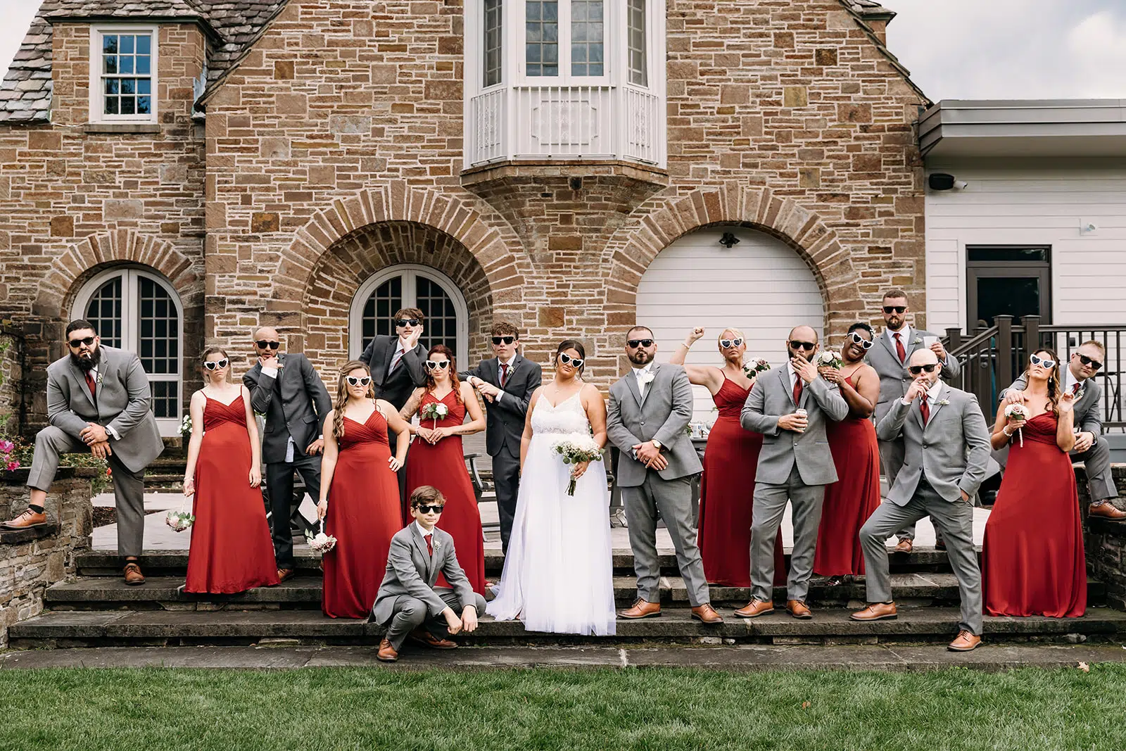 Wedding party posed on the front steps of a brick building, featuring the bride in a white gown, groom and groomsmen in gray suits, and bridesmaids in red dresses.