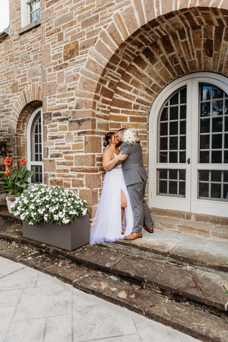 A bride in a white dress and a groom in a gray suit kiss on stone steps beside a brick building with an arched doorway and a planter of white flowers.