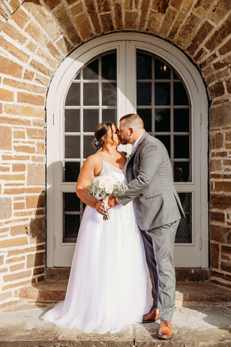 Bride and groom kiss in front of a brick arched doorway, the bride holding a bouquet.