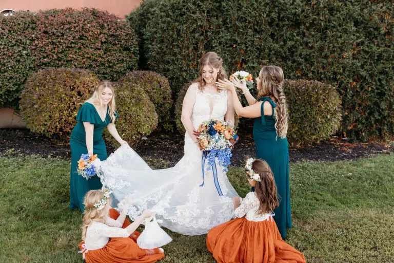 A bride in a white wedding gown stands on a grassy lawn outdoors, assisted by bridesmaids in teal dresses and flower girls in orange dresses.