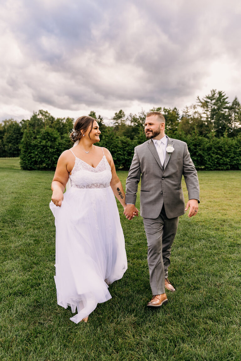 Bride in a white wedding dress and groom in a gray suit walk hand in hand across a grassy field.