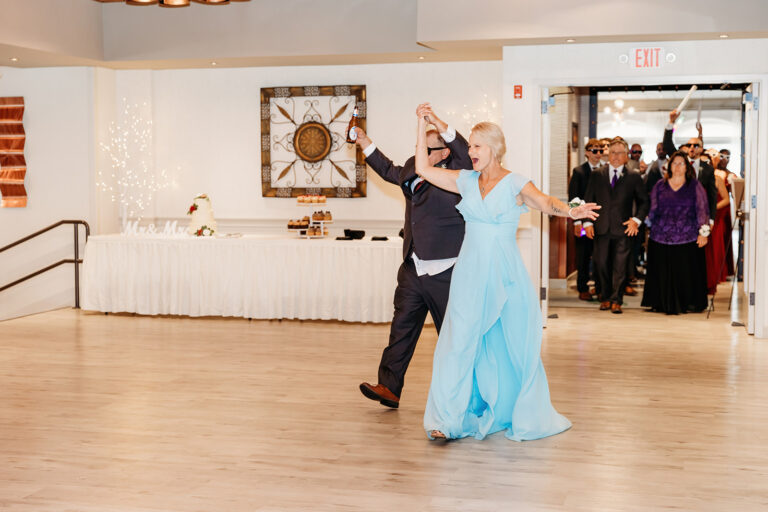 A couple dances on a wooden dance floor at a formal event, with guests watching in the background.