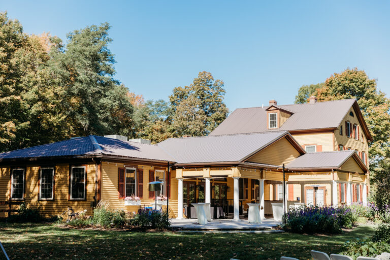 A beige house with a wide front porch and gabled roof sits among trees under a clear blue sky.