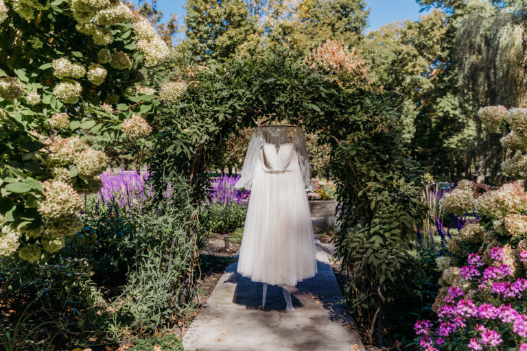A white wedding dress hanging from a leafy arch over a garden path lined with purple flowers.