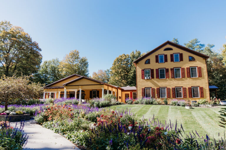 A two-story brick house with red shutters sits beside a single-story yellow building, surrounded by a colorful flower garden and a clear blue sky.