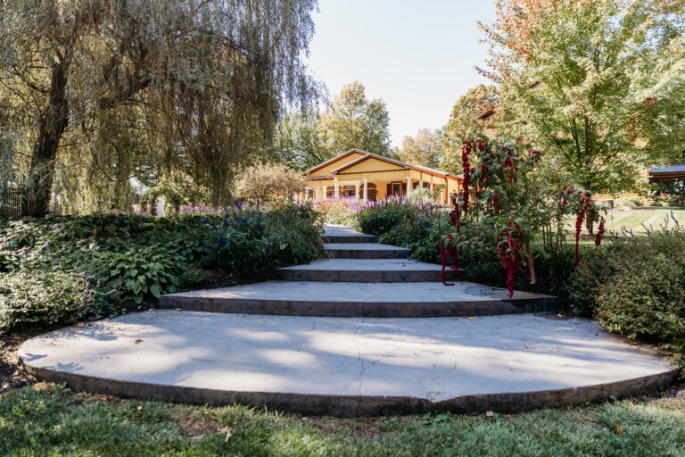 A park garden with a circular concrete platform, steps, and a small wooden pavilion surrounded by trees and shrubs.