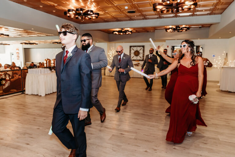 A man in a blue suit and sunglasses walks in front of formally dressed guests dancing at a wedding reception in a ballroom.