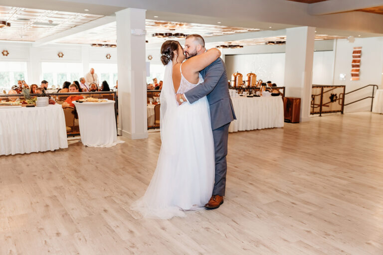 Bride in a white wedding dress and groom in a blue suit share a dance in a bright reception hall with guests seated at long banquet tables in the background.