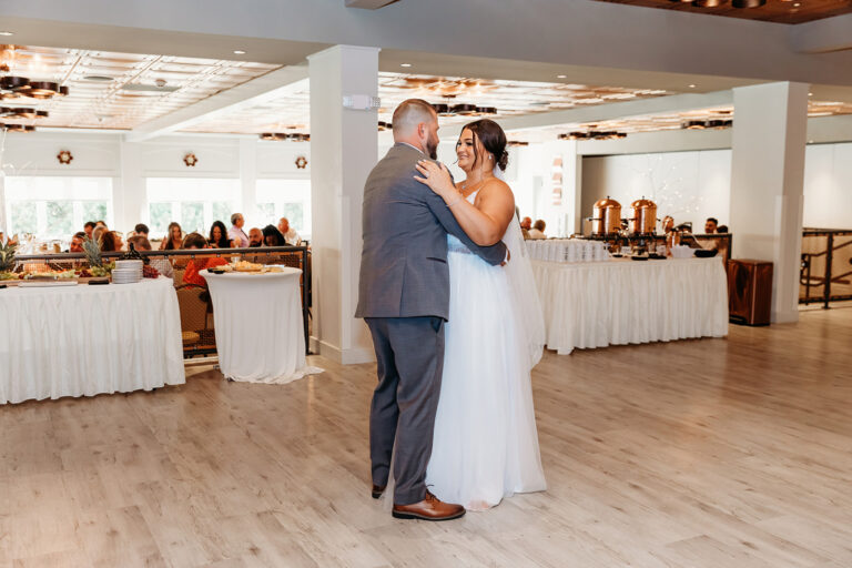 A bride and groom dance together in a wedding reception, with guests seated at long white-clothed tables in the background.