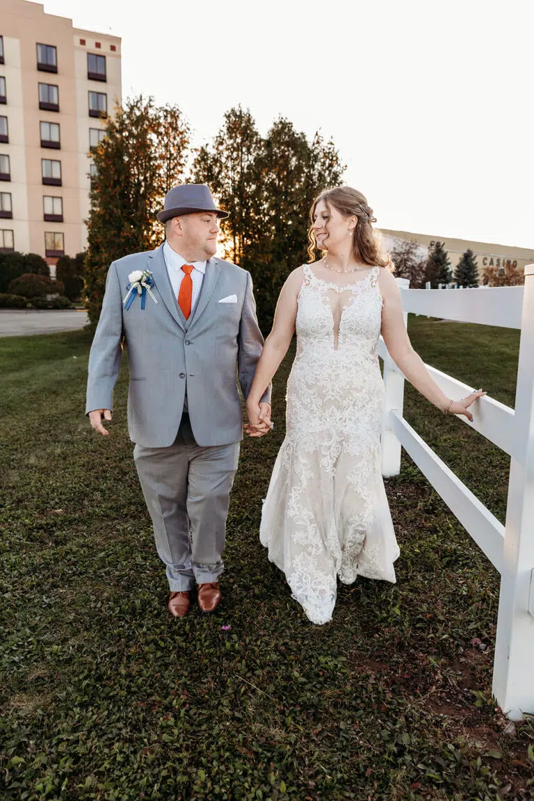 Bride and groom walk hand in hand along a grassy path beside a white fence, with a building and trees in the background.