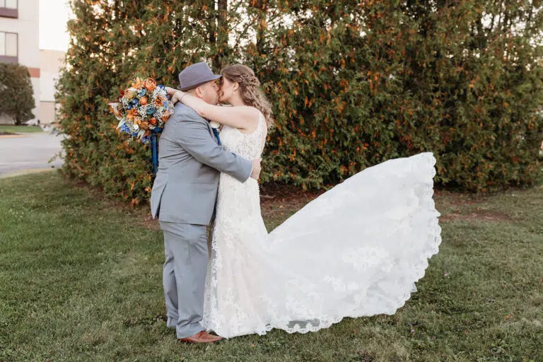 Bride in a white wedding gown and groom in a light gray suit embrace on a grassy lawn outdoors, her train trailing behind.