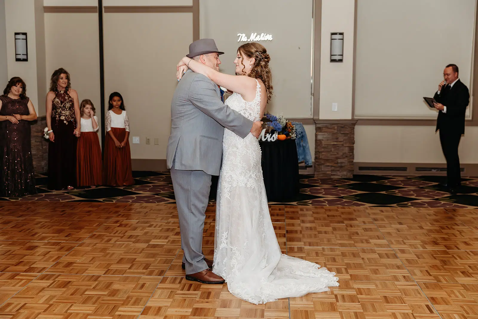 A bride in a white wedding dress dances with a man in a gray suit and hat on a wooden dance floor, as guests watch in the background.