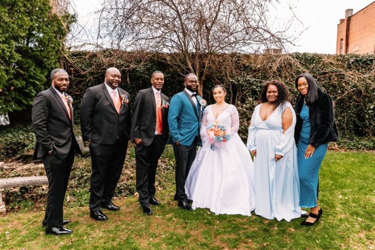 The wedding party poses outdoors on a grassy lawn, with the bride in a white gown holding a bouquet, flanked by bridesmaids in pale blue dresses and groomsmen in suits.