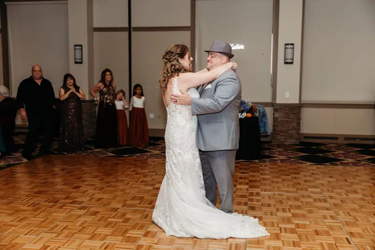 Bride in a white wedding gown dances with a groom in a gray suit on a wood parquet floor at a wedding reception, as guests look on in the background.